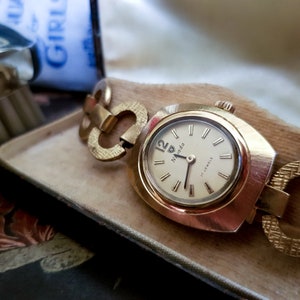 May include: A gold-toned wristwatch with a white face and black hands. The watch has a gold-toned metal band with a unique link design. The watch face has the word "Navada" and "17 Jewels" printed on it.