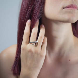 May include: A silver ring with a modern, minimalist design. The ring features a curved, elongated top and a split band that wraps around the finger. The jewelry is worn on a person's hand.