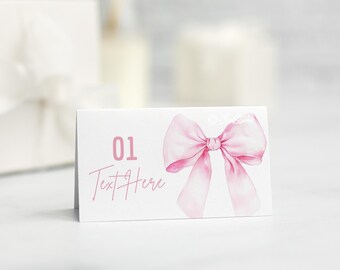 May include: A white place card with a watercolor pink bow illustration. The card features the number "01" and the text "Text Here" in pink. The card is designed for celebrations.