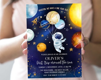 May include: A blue and gold space-themed invitation for a first birthday party. The invitation features an astronaut holding balloons shaped like planets. The text reads "Houston, we have a one year old. Please join us as we celebrate Oliver's first trip around the sun. Sunday | November 14th | 2:00 PM. The Boulevard, 350 West Dillon Road Colorado Springs. RSVP to my mum by 11/12/20XX."