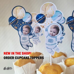 Peut inclure: Un ensemble de d&eacute;corations de cupcakes en forme d'astronautes dans un th&egrave;me bleu, blanc et or. Chaque d&eacute;coration repr&eacute;sente le visage d'un enfant dans une combinaison d'astronaute tenant des ballons avec des plan&egrave;tes et des &eacute;toiles. Le texte "NEW IN THE SHOP! ORDER CUPCAKE TOPPERS" est en bas de l'image.