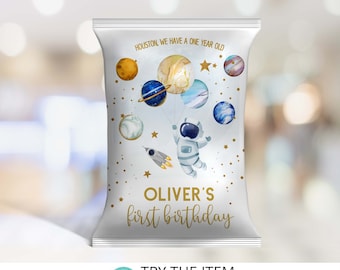 May include: A white chip bag with a gold astronaut holding blue and gold planets. The text "HOUSTON, WE HAVE A ONE YEAR OLD" and "OLIVER'S first birthday" is printed on the bag.