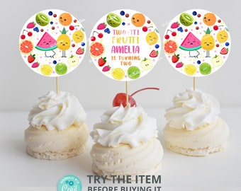 May include: Two cupcake toppers with a white background and colorful fruit designs. The toppers feature a watermelon, pineapple, and other fruits. The text on the toppers reads "TWO-TTI FRUTTI AMELIA IS TURNING TWO".