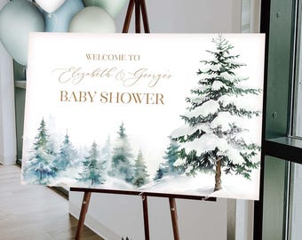 May include: A welcome sign for a baby shower, featuring a winter forest scene with snow-covered trees. The sign reads "Welcome to Elizabeth & George's Baby Shower" in gold lettering. The sign is displayed on a wooden easel, with balloons in the background.