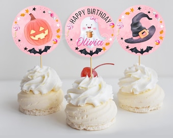 Op de afbeelding: Drie Halloween-cupcakes met roze toppers. De toppers tonen een pompoen, een spookje met een strik en een heksenhoed. Elke cupcake is bedekt met witte frosting. De spooktopper zegt "Happy Birthday Olivia".