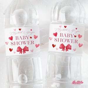 Puede incluir: Dos botellas de agua de plástico transparente con etiquetas rojas y blancas. Las etiquetas tienen un diseño de acuarela de corazones y un lazo. El texto en las etiquetas dice "Elizabeth & George Baby Shower".