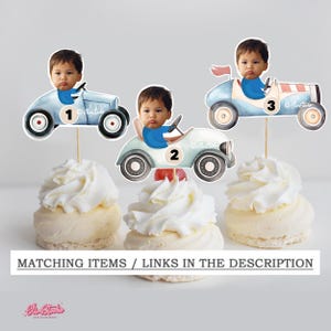 Op de afbeelding: Cupcake toppers met een babygezicht in lichtblauwe oldtimer raceauto's. De auto's hebben zwarte wielen en de nummers 1, 2 en 3. De cupcakes zijn bedekt met witte glazuur. De tekst onderaan luidt "MATCHING ITEMS / LINKS IN THE DESCRIPTION."