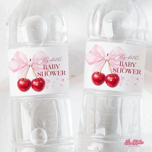 May include: Two clear plastic water bottles with labels that read "Elizabeth's BABY SHOWER." The labels feature a pink bow, two red cherries, and pink stars. The bottles are filled with water and have a white cap.