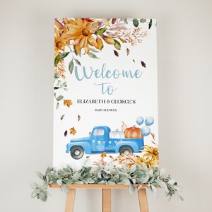Op de afbeelding: Welkom bij de babyshower van Elizabeth & George bord met een blauwe vrachtwagen die pompoenen en ballonnen vervoert, omringd door herfstbladeren en bloemen.