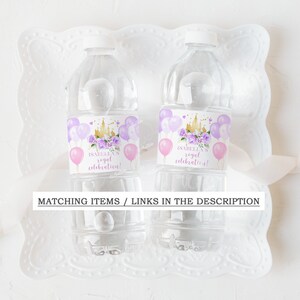 Op de afbeelding: Twee doorzichtige plastic waterflessen met paarse en roze etiketten. De etiketten tonen een gouden kasteel, paarse bloemen en roze ballonnen. De tekst op de etiketten luidt "Isabella's royal celebration!"