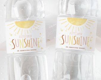 Könnte beinhalten: Zwei durchsichtige Plastikwasserflaschen mit Etiketten, auf denen "Ein kleiner Sonnenstrahl kommt bald!" steht. Die Etiketten zeigen eine gelbe Sonne mit einem rosa-orangenen Farbverlauf, umgeben von goldenen Sternen.