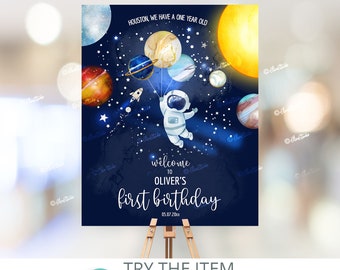 May include: A blue and white astronaut holding balloons shaped like planets against a dark blue background with stars. The text "Houston, we have a one year old" and "Welcome to Oliver's first birthday 05.07.20xx" is printed on the poster.