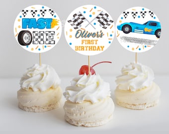 May include: Three round cupcake toppers with a racing car theme. The toppers feature a blue race car, checkered flags, and the text "Fast One" and "Oliver's First Birthday".