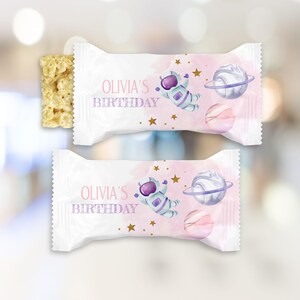 May include: Two white candy bar wrappers with a pink and purple space-themed design. The wrappers feature an astronaut, planets, and the text "OLIVIA'S BIRTHDAY". A partially visible rice crispy treat is on the left.