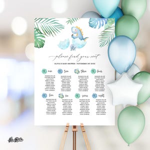 May include: A baby shower seating chart on a wooden easel. The sign features a watercolor illustration of a dinosaur hatching from an egg, with the text "please find your seat." The names are organized by table number, with balloons in the background.