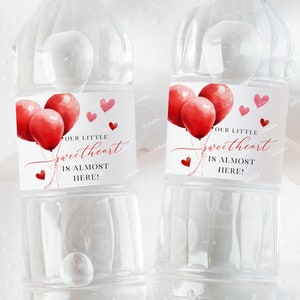 Puede incluir: Dos botellas de agua de plástico transparente con etiquetas que dicen "Nuestro pequeño amorcito está casi aquí!" con globos rojos de acuarela y corazones.