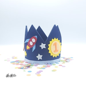 Puede incluir: Corona de fieltro azul con un cohete, estrellas y un sol amarillo con el número 1. La corona está rodeada de confeti de colores.