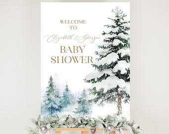 May include: A baby shower welcome sign on an easel. The sign features a winter forest scene with snow-covered evergreen trees and the words "Welcome to Elizabeth & Georges Baby Shower" in gold lettering. Decorative garland and ornaments adorn the easel.