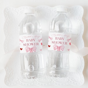 Puede incluir: Dos botellas de agua de plástico transparente con tapones blancos. Cada botella tiene una etiqueta que dice "Elizabeth & George BABY SHOWER" con lazos y corazones rosas. Las botellas están sobre una bandeja decorativa blanca.