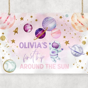 Op de afbeelding: Een roze en witte ruimte-thema feestbanner met gouden sterren en planeten. De banner luidt "Olivia's first trip around the sun" en toont een cartoon-astronaut.