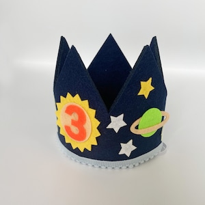 Pode incluir: Uma coroa de feltro azul com um sol amarelo, o número 3 laranja, estrelas brancas e um planeta verde com um anel marrom. A coroa tem uma borda branca e azul.