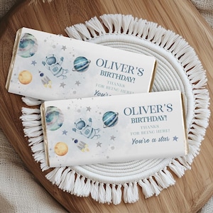 May include: Two chocolate bar wrappers with a space theme. The wrappers feature illustrations of planets, astronauts, rockets, and stars. Text on the wrappers reads "OLIVER'S BIRTHDAY! THANKS FOR BEING HERE! You're a star."
