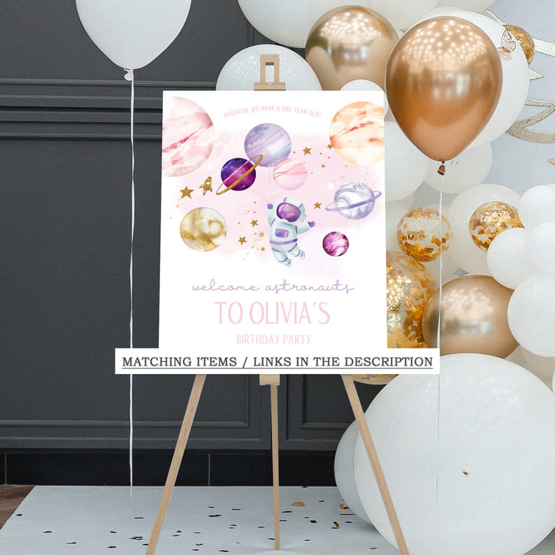 May include: A pink and white welcome sign with a cartoon astronaut and planets. The sign says "Houston, we have a one year old! Welcome astronauts to Olivia's Birthday Party".