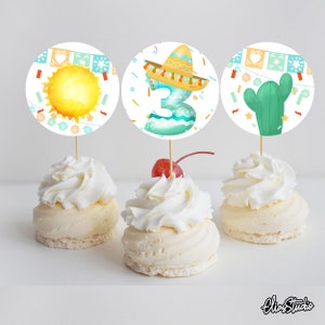 May include: Three cupcake toppers with a fiesta theme. The toppers feature a yellow sun, a blue number three with a sombrero, and a green cactus.