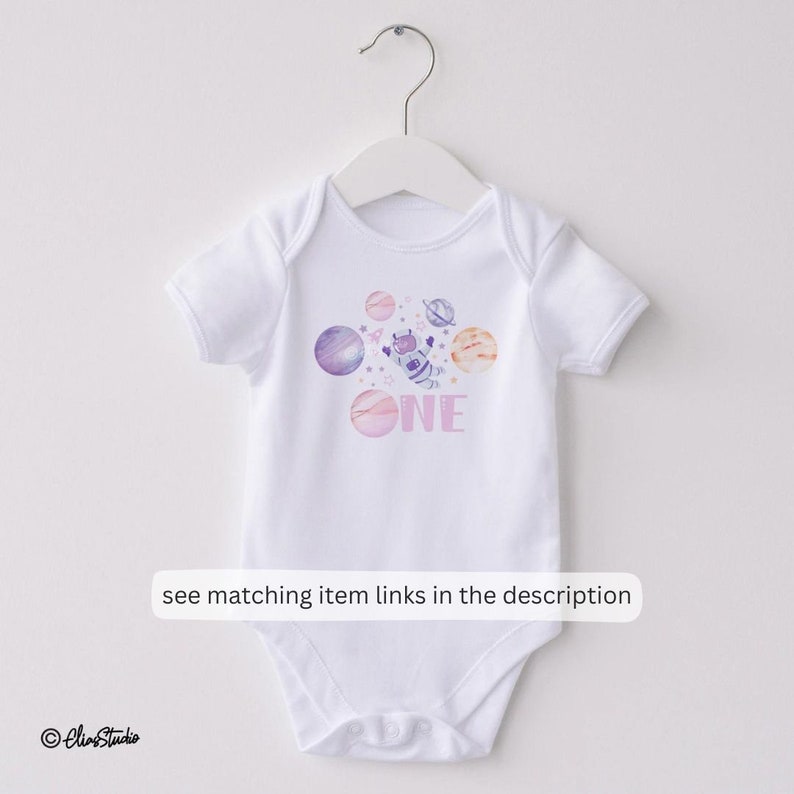 May include: White baby onesie with a pink, purple, and blue watercolor design of planets, stars, and an astronaut. The onesie says "ONE" in purple letters.