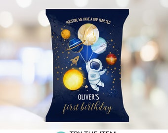 May include: A blue and gold astronaut-themed party favor bag with a cartoon astronaut holding balloons shaped like planets. The text "HOUSTON, WE HAVE A ONE YEAR OLD" and "OLIVER'S first birthday" is printed on the bag.