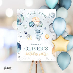May include: A birthday party sign with an astronaut illustration holding planets, a rocket, and the text "5,4,3,2...ONE!" and "Welcome to Oliver's birthday party!" in blue and gold. The sign is on a wooden easel, with blue and gold balloons.