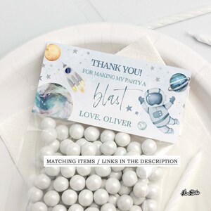 May include: A blue and white thank you favor bag with a space theme. The bag features an astronaut, planets, stars, and a rocket. The text on the bag reads "Thank you! For making my party a blast. Love, Oliver."