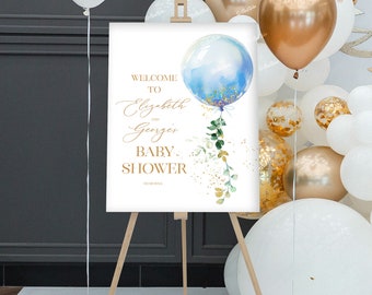 Puede incluir: Bienvenido a la fiesta de bienvenida al bebé de Elizabeth y George, cartel con una ilustración de un globo azul y dorado. El cartel está en un caballete con globos blancos y dorados en el fondo.