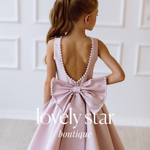 May include: A light pink sleeveless dress with a large bow in the back. The dress is decorated with a row of pearls around the neckline and the bow. The dress is made of a satin-like fabric. The text "lovely star boutique" is printed on the dress.