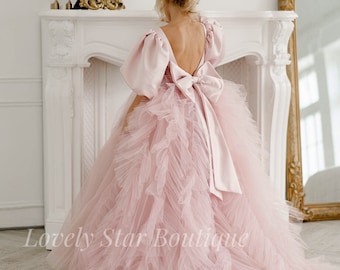May include: A light pink formal gown with a voluminous skirt and a large bow at the back. The dress features puffy sleeves and a low back. The text "Lovely Star Boutique" is visible on the bottom left of the image.