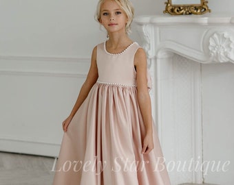 May include: A young girl wearing a blush pink satin dress with a white pearl trim. The dress has a full skirt and a sleeveless bodice. The girl is standing in front of a white fireplace with a gold mirror above it.