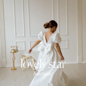 K&ouml;nnte beinhalten: Ein junges M&auml;dchen tr&auml;gt ein wei&szlig;es Satin-Kleid mit einer gro&szlig;en Schleife im R&uuml;cken. Das Kleid hat einen V-Ausschnitt und Puff&auml;rmel. Das M&auml;dchen steht in einem wei&szlig;en Raum mit einem goldenen Telefon auf einem St&auml;nder im Hintergrund. Der Text "lovely star boutique" ist im Bild sichtbar.