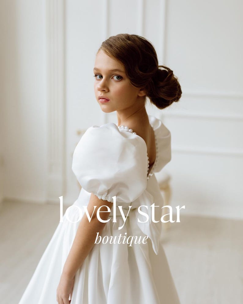 K&ouml;nnte beinhalten: Ein junges M&auml;dchen tr&auml;gt ein wei&szlig;es Kleid mit Puff&auml;rmeln und Perlenbesatz. Das Kleid hat einen vollen Rock und eine Schleife an der Taille. Das M&auml;dchen schaut &uuml;ber die Schulter und l&auml;chelt. Der Text "lovely star boutique" ist auf das Kleid gedruckt.