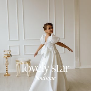 K&ouml;nnte beinhalten: Ein junges M&auml;dchen tr&auml;gt ein wei&szlig;es Kleid mit einer gro&szlig;en Schleife im R&uuml;cken. Das Kleid hat Puff&auml;rmel und einen V-Ausschnitt. Das M&auml;dchen steht in einem wei&szlig;en Raum mit einem wei&szlig;en Hocker und einem goldenen Telefonst&auml;nder. Der Text "lovely star boutique" ist im Bild sichtbar.