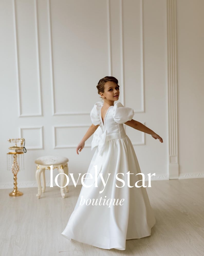 K&ouml;nnte beinhalten: Ein junges M&auml;dchen tr&auml;gt ein wei&szlig;es Kleid mit einer gro&szlig;en Schleife im R&uuml;cken. Das Kleid hat Puff&auml;rmel und einen V-Ausschnitt. Das M&auml;dchen steht in einem wei&szlig;en Raum mit einem wei&szlig;en Hocker und einem goldenen Telefonst&auml;nder. Der Text "lovely star boutique" ist im Bild sichtbar.