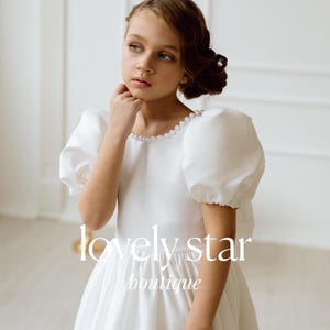 K&ouml;nnte beinhalten: Ein junges M&auml;dchen tr&auml;gt ein wei&szlig;es Kleid mit Puff&auml;rmeln und Perlenbesatz. Das Kleid hat einen vollen Rock und ist perfekt f&uuml;r einen besonderen Anlass. Das M&auml;dchen schaut zur Seite und hat einen nachdenklichen Ausdruck im Gesicht. Der Text "lovely star boutique" ist unten im Bild sichtbar.