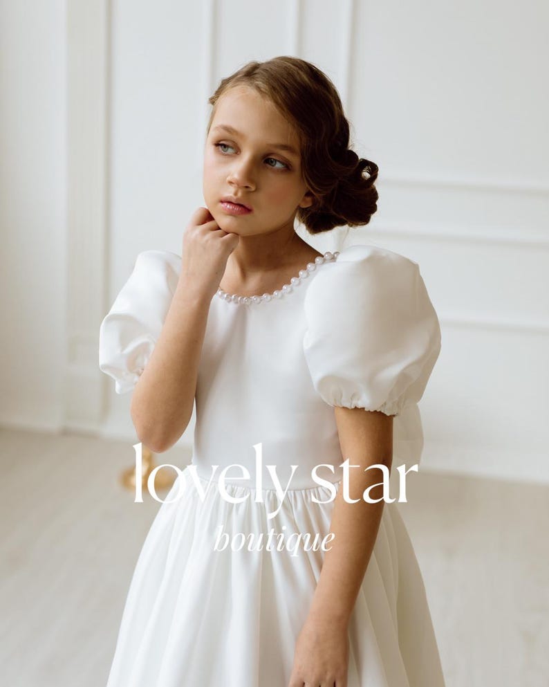 K&ouml;nnte beinhalten: Ein junges M&auml;dchen tr&auml;gt ein wei&szlig;es Kleid mit Puff&auml;rmeln und Perlenbesatz. Das Kleid hat einen vollen Rock und ist perfekt f&uuml;r einen besonderen Anlass. Das M&auml;dchen schaut zur Seite und hat einen nachdenklichen Ausdruck im Gesicht. Der Text "lovely star boutique" ist unten im Bild sichtbar.