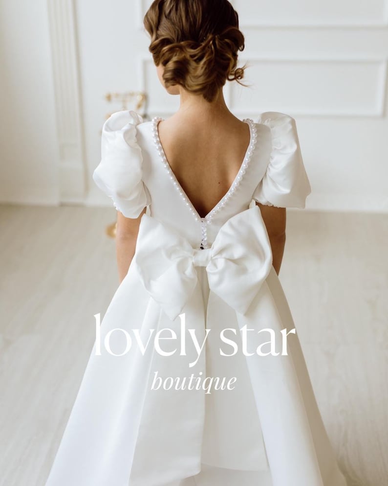 K&ouml;nnte beinhalten: Wei&szlig;es Satin-Kleid mit einer gro&szlig;en Schleife im R&uuml;cken und einem Perlenbesatz am Ausschnitt. Das Kleid hat Puff&auml;rmel und einen V-f&ouml;rmigen R&uuml;cken. Das Kleid ist von der "lovely star boutique".