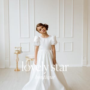 K&ouml;nnte beinhalten: Ein junges M&auml;dchen tr&auml;gt ein wei&szlig;es Kleid mit Puff&auml;rmeln und Perlenbesatz. Das Kleid hat einen vollen Rock und ist perfekt f&uuml;r einen besonderen Anlass. Das M&auml;dchen steht in einem wei&szlig;en Raum mit einem goldenen Dekorst&auml;nder hinter ihr. Der Text "lovely star boutique" ist auf das Bild gedruckt.