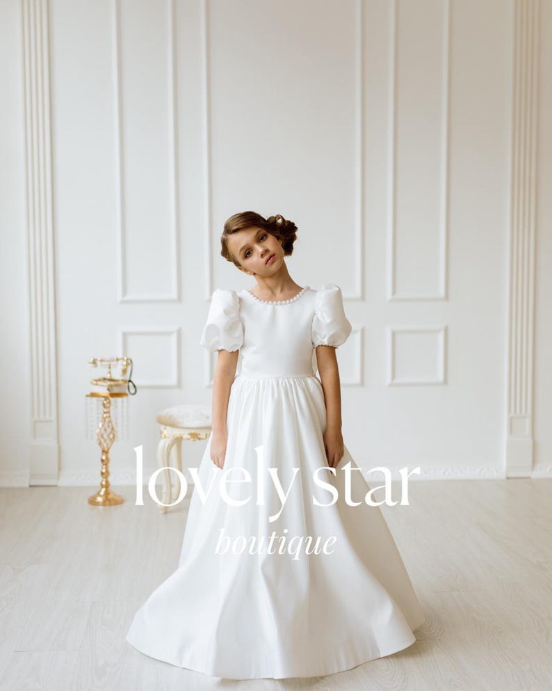K&ouml;nnte beinhalten: Ein junges M&auml;dchen tr&auml;gt ein wei&szlig;es Kleid mit Puff&auml;rmeln und Perlenbesatz. Das Kleid hat einen vollen Rock und ist perfekt f&uuml;r einen besonderen Anlass. Das M&auml;dchen steht in einem wei&szlig;en Raum mit einem goldenen Dekorst&auml;nder hinter ihr. Der Text "lovely star boutique" ist auf das Bild gedruckt.