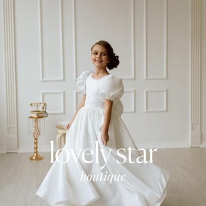 K&ouml;nnte beinhalten: Ein junges M&auml;dchen tr&auml;gt ein wei&szlig;es Kleid mit Puff&auml;rmeln und einem vollen Rock. Das Kleid hat einen Perlensaum um den Ausschnitt. Das M&auml;dchen steht in einem wei&szlig;en Raum mit einem goldenen Telefon auf einem St&auml;nder hinter ihr. Der Text "lovely star boutique" ist unten im Bild gedruckt.