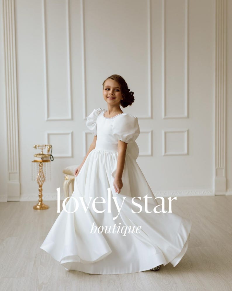 K&ouml;nnte beinhalten: Ein junges M&auml;dchen tr&auml;gt ein wei&szlig;es Kleid mit Puff&auml;rmeln und einem vollen Rock. Das Kleid hat einen Perlensaum um den Ausschnitt. Das M&auml;dchen steht in einem wei&szlig;en Raum mit einem goldenen Telefon auf einem St&auml;nder hinter ihr. Der Text "lovely star boutique" ist unten im Bild gedruckt.