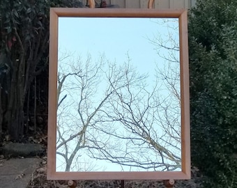 May include: A rectangular wooden framed mirror reflecting a blue sky and bare tree branches. The mirror is mounted on a wooden easel. Two decorative wooden pieces are on top of the frame.