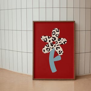 Op de afbeelding: Een ingelijst afdruk van een rode achtergrond met een grote, gestileerde bloem. De bloem is wit en zwart met een luipaardprint. De steel van de bloem is lichtblauw.