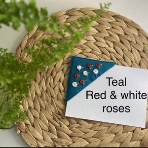May include: Teal fabric bookmark with red and white embroidered roses. The bookmark is on a woven placemat. Teal Red & white roses
